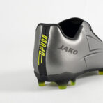 Close-up of a silver JAKO soccer boot heel with a black stripe, lime-yellow studs, and branding visible on the side.