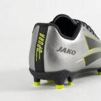 Rear view of a silver soccer cleat with neon yellow accents and the Jako logo on the heel area.